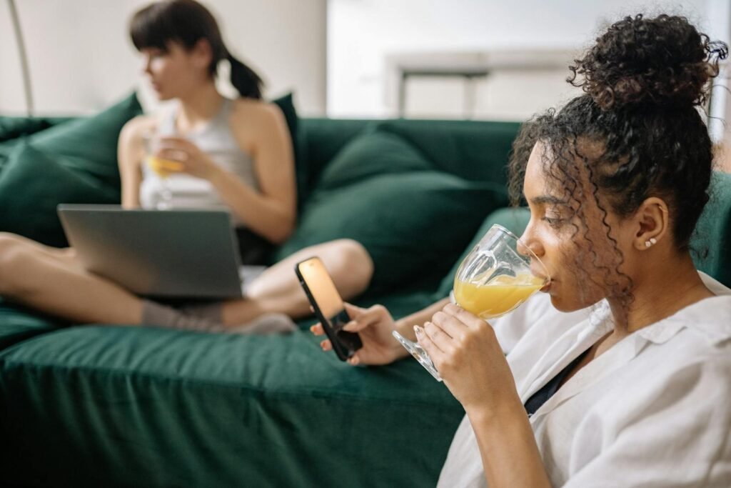 Two young women enjoying drinks at home, one with a phone and another with a laptop.